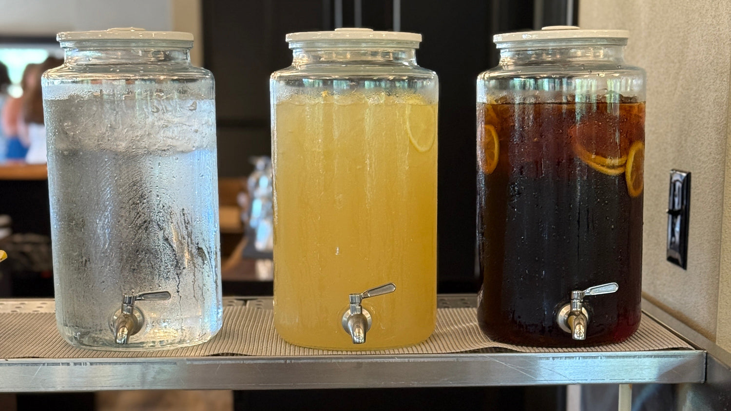 Three large glass beverage dispensers on a stainless steel counter, filled with ice water, lemonade, and iced tea, are included in our beverage package.